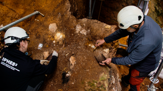 Two archaeologists dig in reddish-brown dirt to expose small fragments of fossil bone