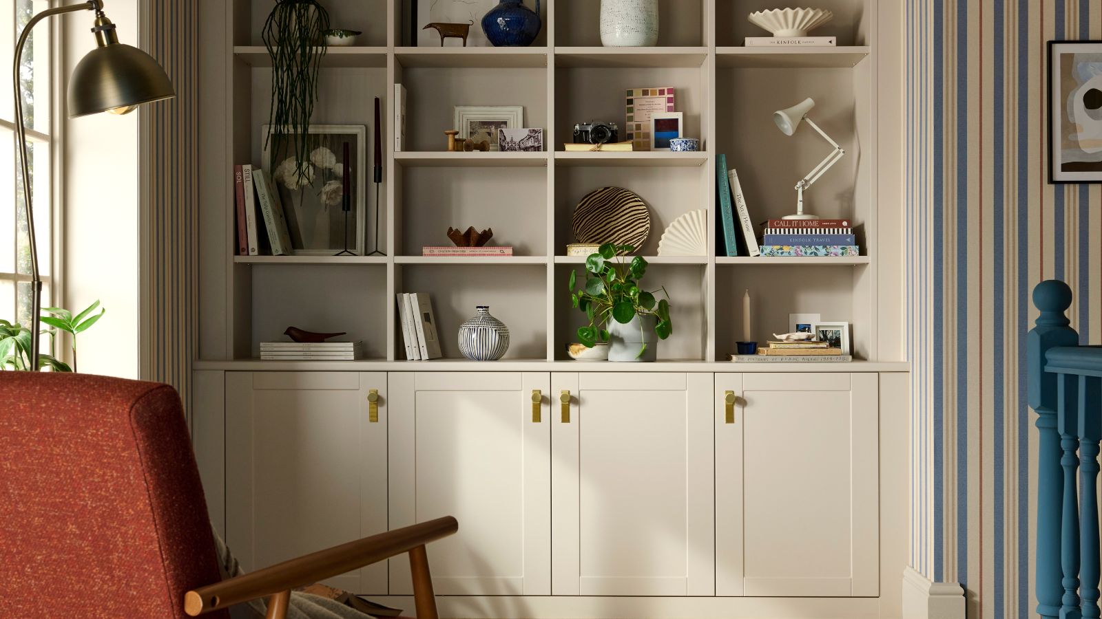 A beautifully styled landing features a built-in shelving unit painted in a warm neutral shade. The upper shelves are neatly arranged with books, ceramics, framed photos, and a leafy plant, creating a balanced yet homely look.
