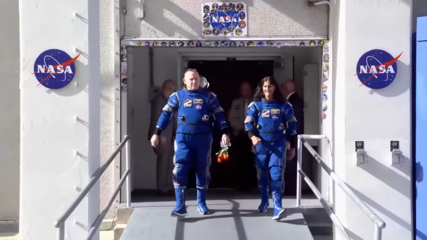 Dos astronautas con trajes espaciales azules caminan uno al lado del otro y sonríen.