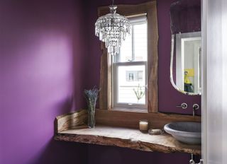 A bathroom with purple walls, and a small chandelier hung above the wooden counter