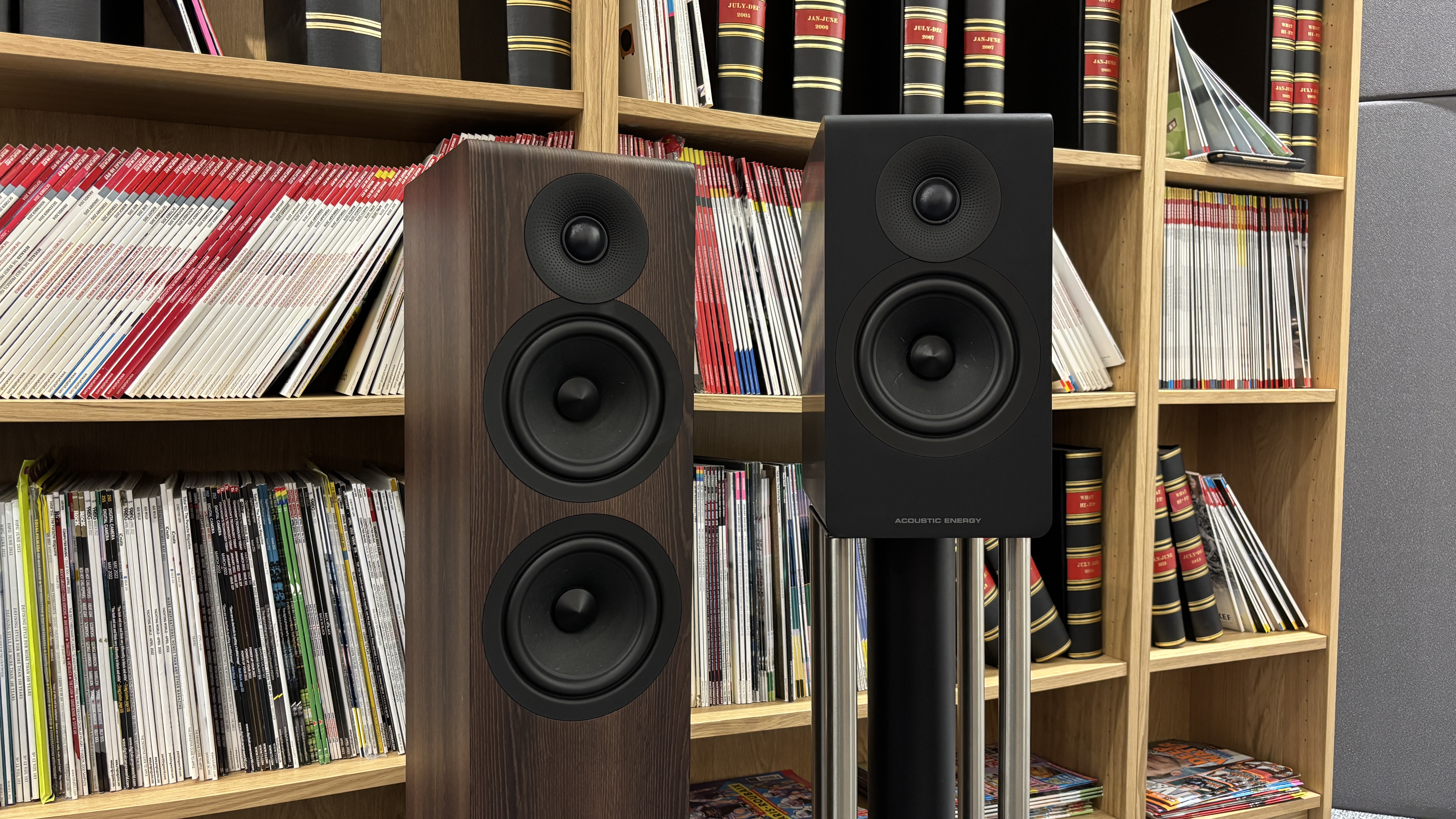 Acoustic Energy AE309 Mk2 and AE300 Mk2 standing next to each other in front of bookshelves filled with magazines