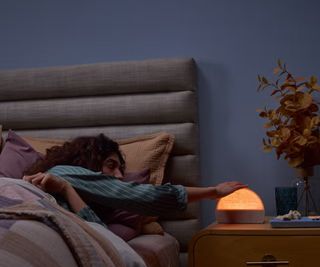 A woman in bed switching on the Hatch Restore 2 Sunrise Alarm Clock on a nightstand against a blue wall.