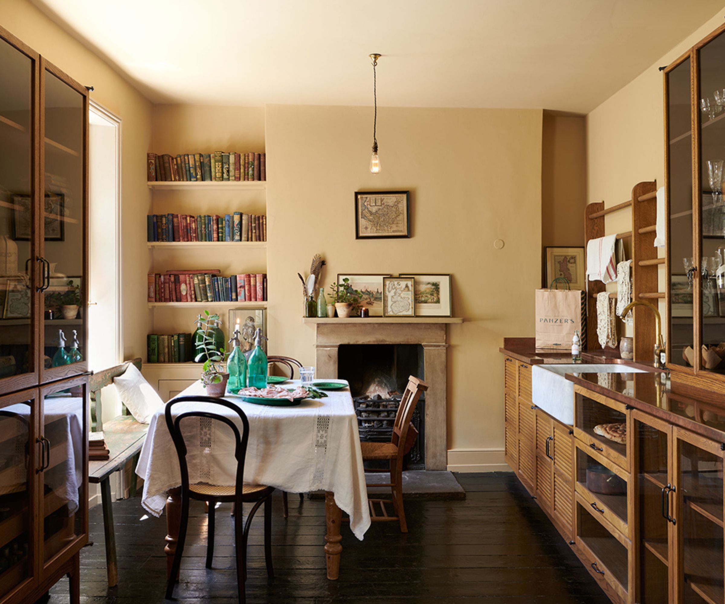 A pale yellow kitchen with wooden freestanding cabinets, a wall of book shelves, a traditional fireplace, and a large dining table covered in a white linen tablecloth