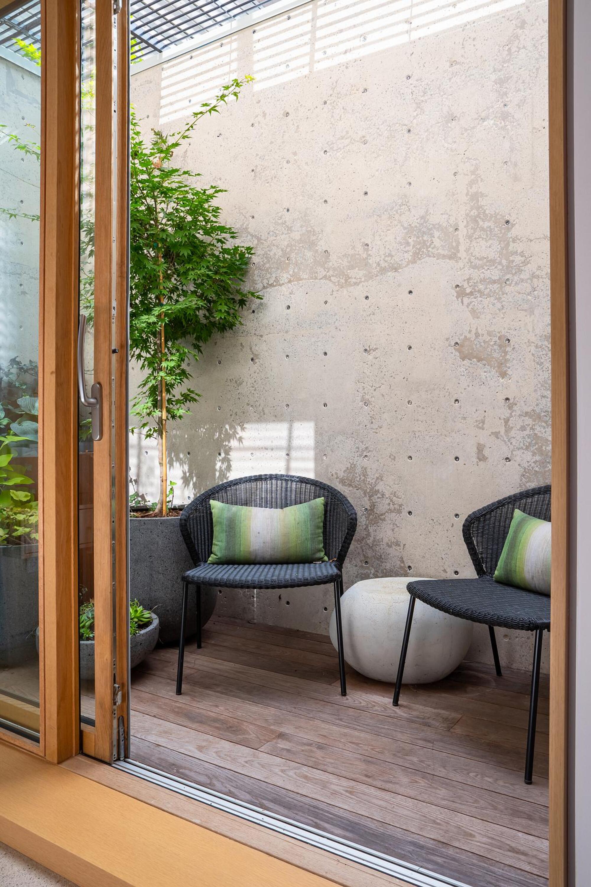 small courtyard garden off a bedroom with concrete wall, potted tree, two woven armchairs with outdoor cushions on them and a round cement side table between them