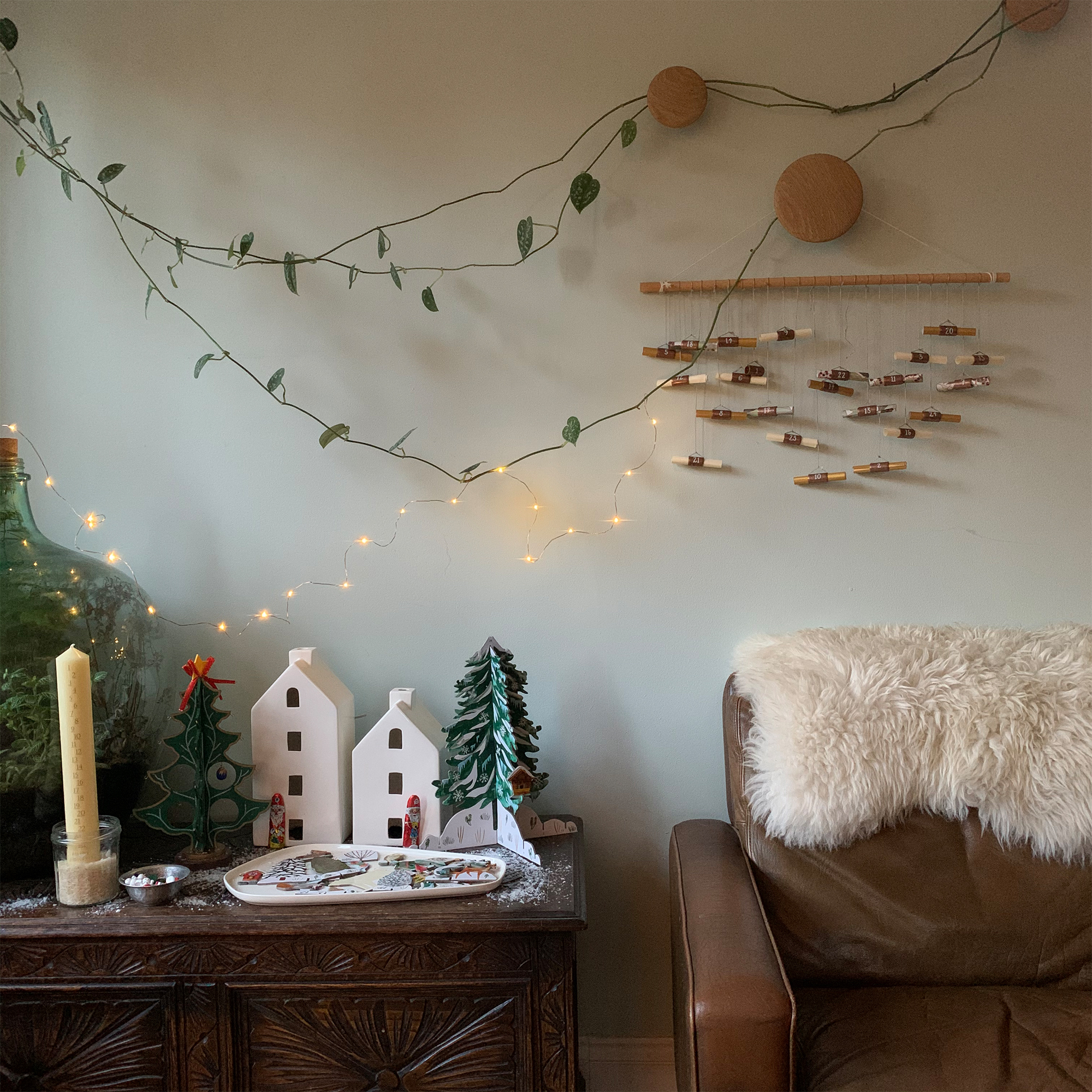 Advent scene with pale green wall and wall-hung activity advent calendar, string of wire fairy lights and a tabletop display of ceramic houses and advent trees