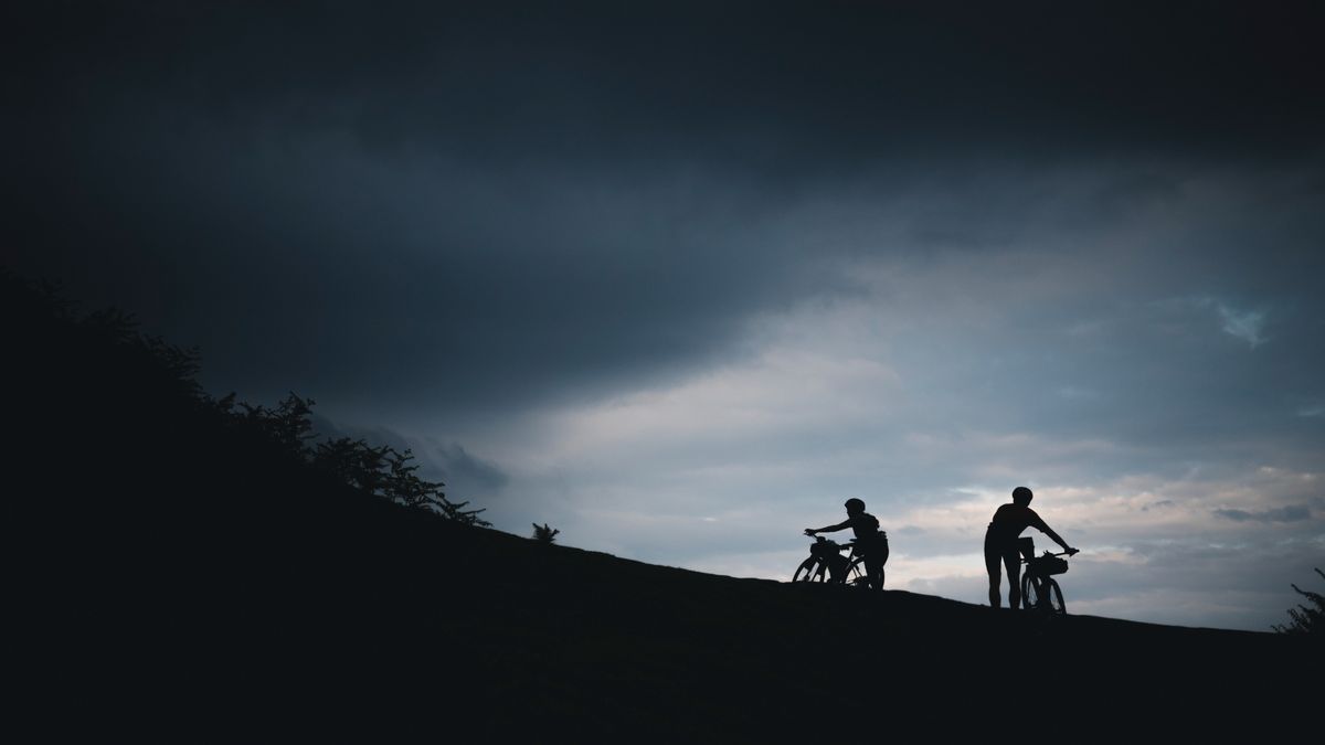 Two cyclists push their bikes up a hill in silhouetted against a dark sky