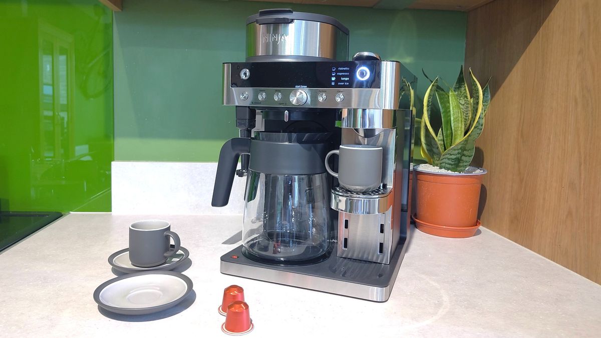 Ninja Prestige DualBrew System coffee maker on kitchen counter with plant, coffee cups with saucers, and coffee pods