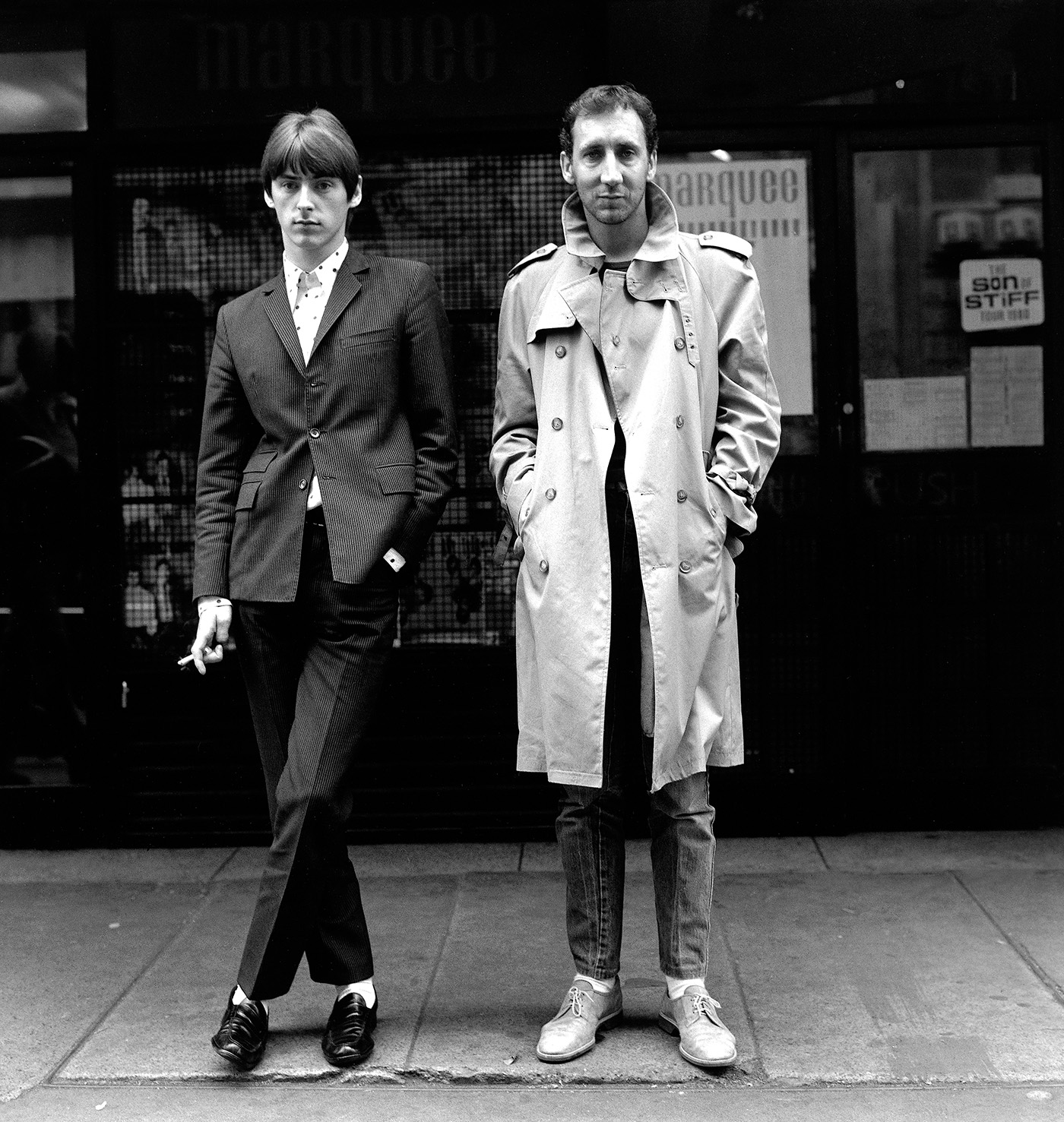 Black and white photograph of Paul Weller and Pete Townshend standing on a London pavement outside the Marquee Club, one in a pinstripe suit and the other in a trench coat.