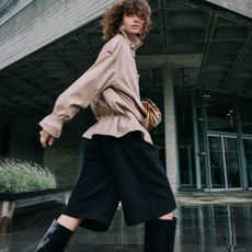 An upward shot of a model wearing black knee high boots, black knee length shorts, and a tan jacket cinched at the waist
