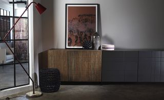 The space, quite dark and grey focusses on a dark wood wall cabinet with a red and black painting on it. In the foreground is a large floor lamp.