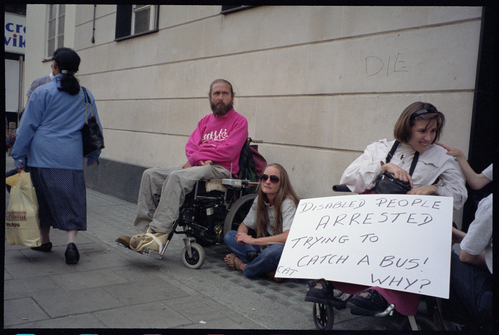Disability rights protesters, including wheelchair users, gather outside a London building with placards, one reading "People with disabilities unite against your need to segregate us."