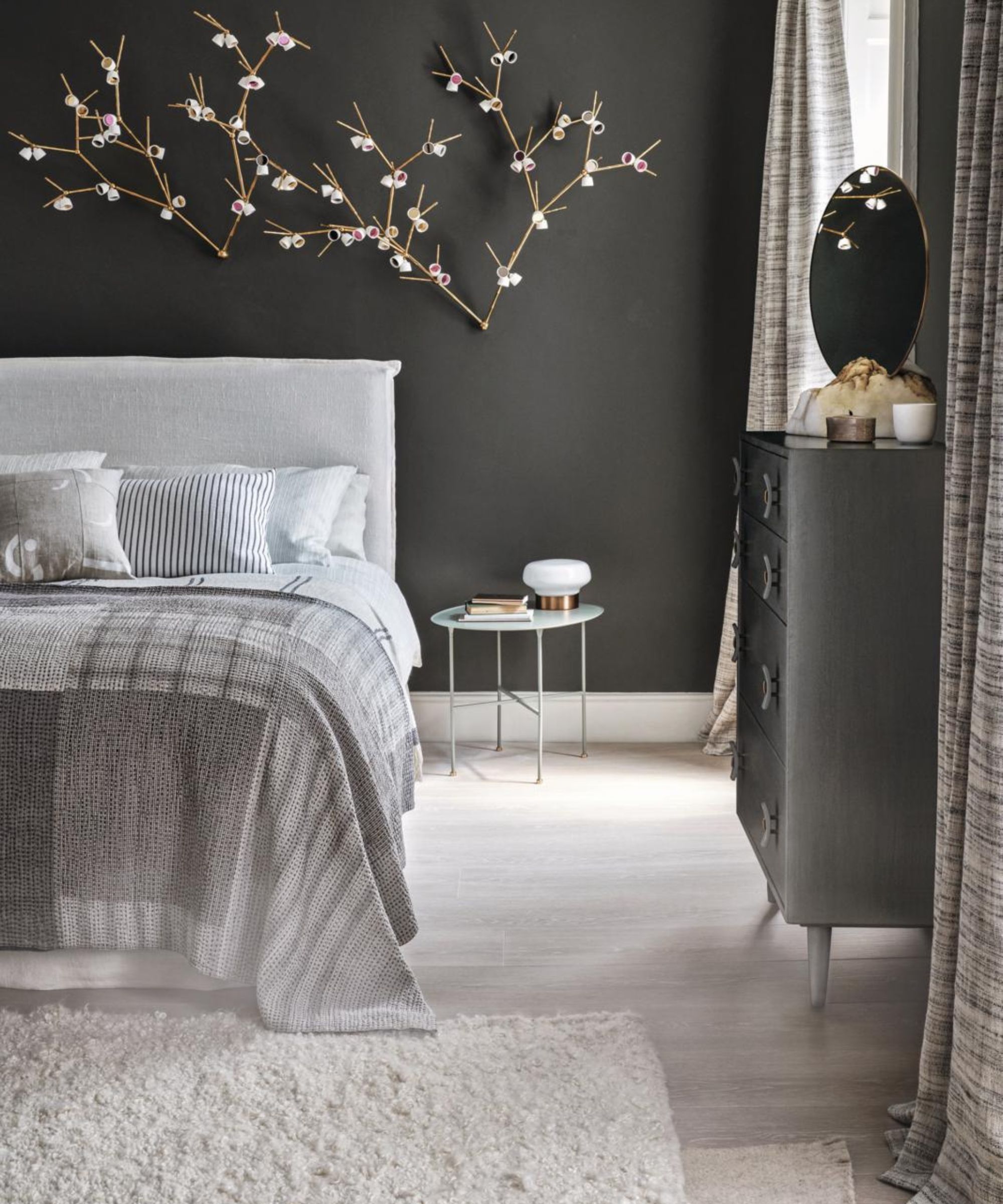 A dark gray bedroom with a large double bed with neutral bedding, wooden flooring, a large white shaggy rug, and a gray chest of drawers to the right with a circular gold mirror. On the dark gray wall behind the bed is a statement artwork piece of cherry blossoms.