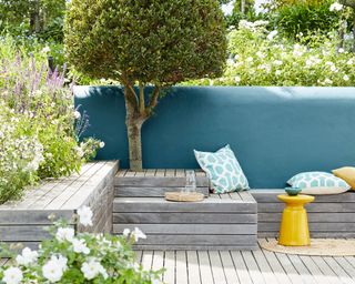 Small garden decking ideas example with levels of built-in seating and steps leading up to a small tree, with blue wall and yellow side table.