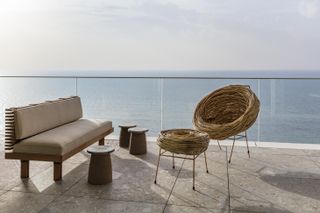 balcony with a brown bench seat, stools and wicker chair and footrest, with clear glass walls showing the sea view