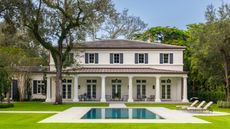 an exterior shot of a large white home with a pillared covered patio area out the back looking over a grass area and swimming pool surrounded by trees