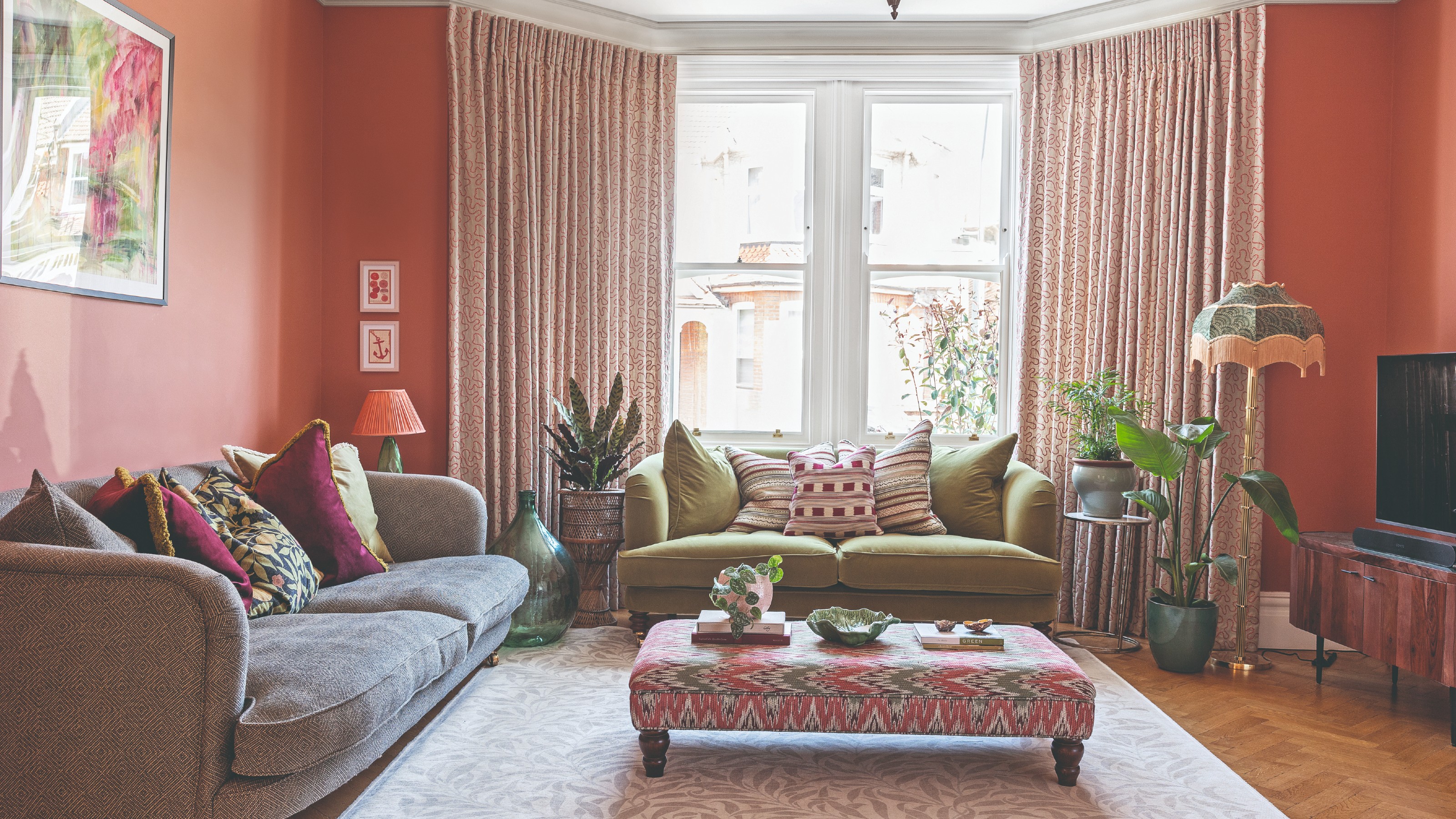 A coral-painted hallway with patterned curtains, a grey and an olive green sofa and an ottoman used as a coffee table