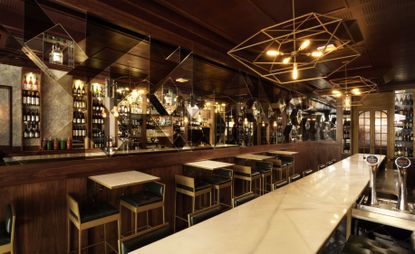 A bar with a long light marble counter, tables, chairs, wall mirrors and wooden walls.