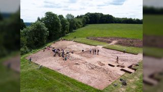 Overview of a Neolithic burial site at Dorstone Hill in the west of England. Here we see a large rectangle that has been dug up in a grassy field. There are a lot of people working on the site.