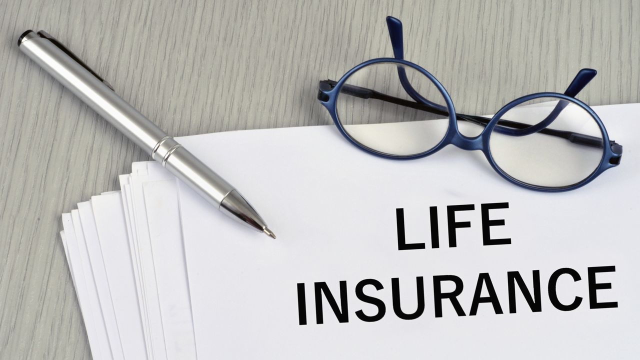 The words life insurance on white paper with a pen and eyeglasses on top.