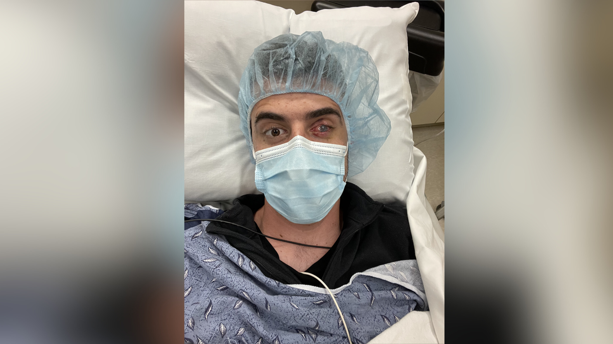 A photo of Nick as he is sat in a hospital bed following surgery. He is wearing a blue hair net and a blue face mask. 