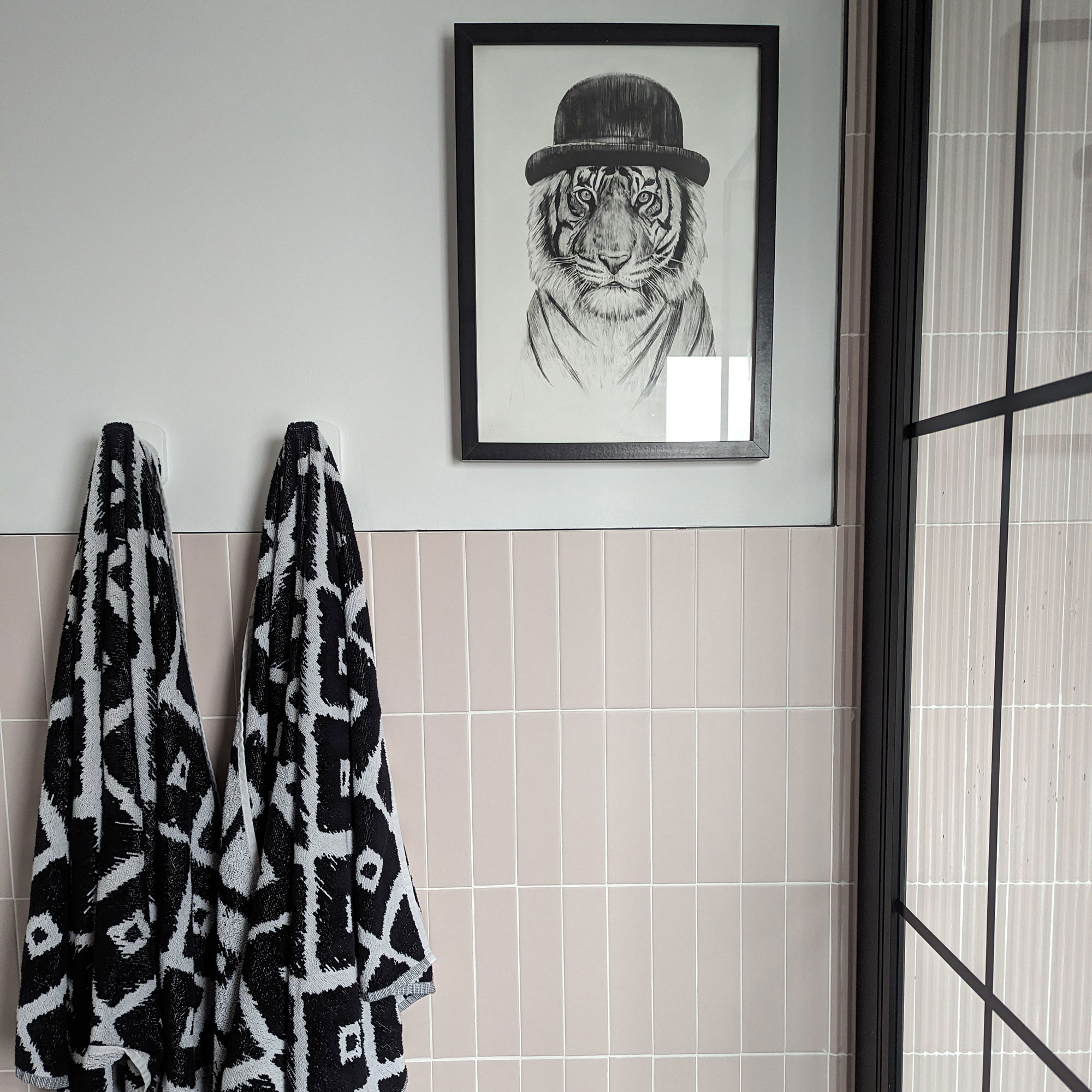 Pink tiled bathroom wall with monochrome patterned towels hanging up and artwork of tiger wearing bowler hat