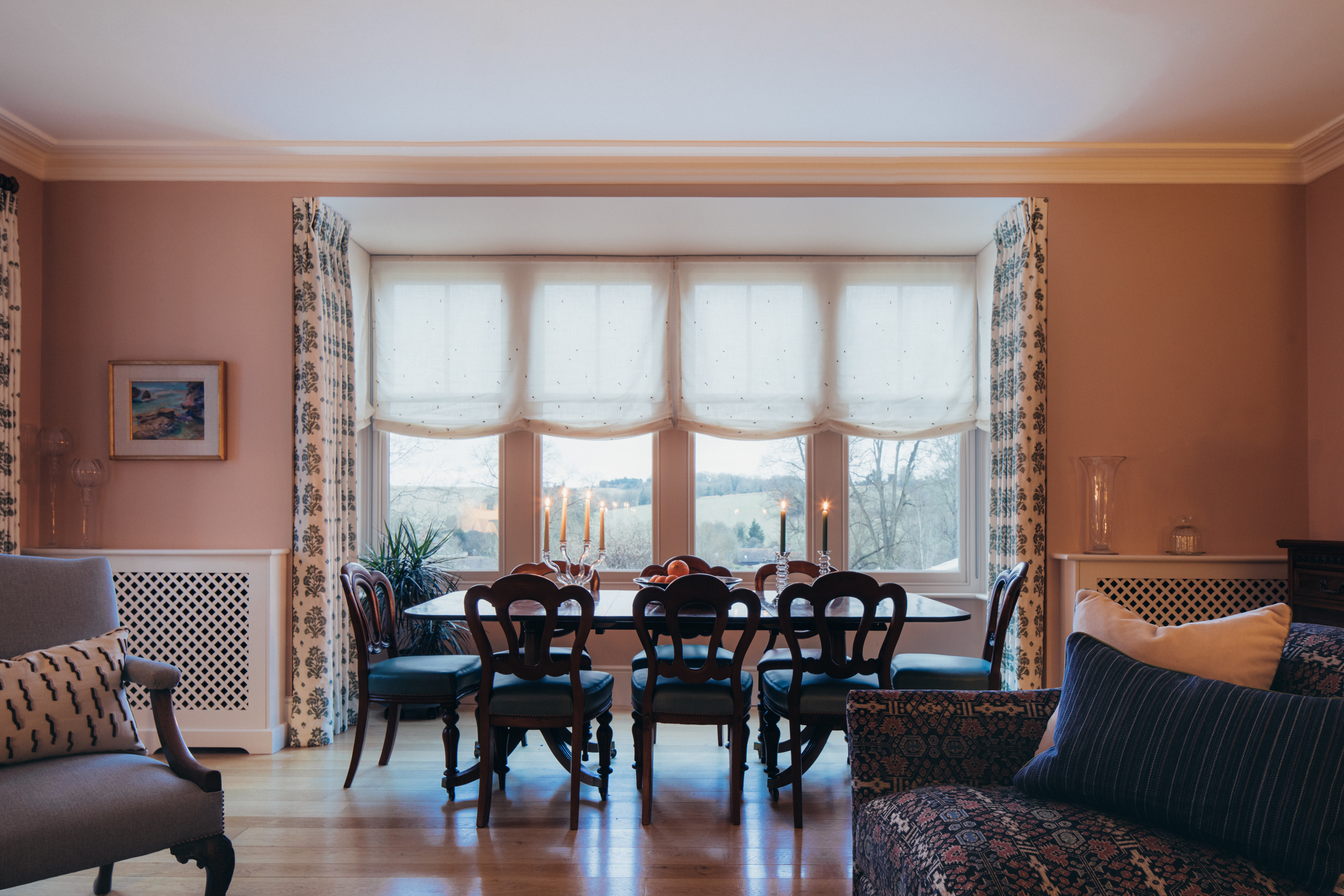 a dining table sits in a peach living area in front of a large alcove window with two lit candelabras on the table and two white radiator covers sits either side of the window and dining area