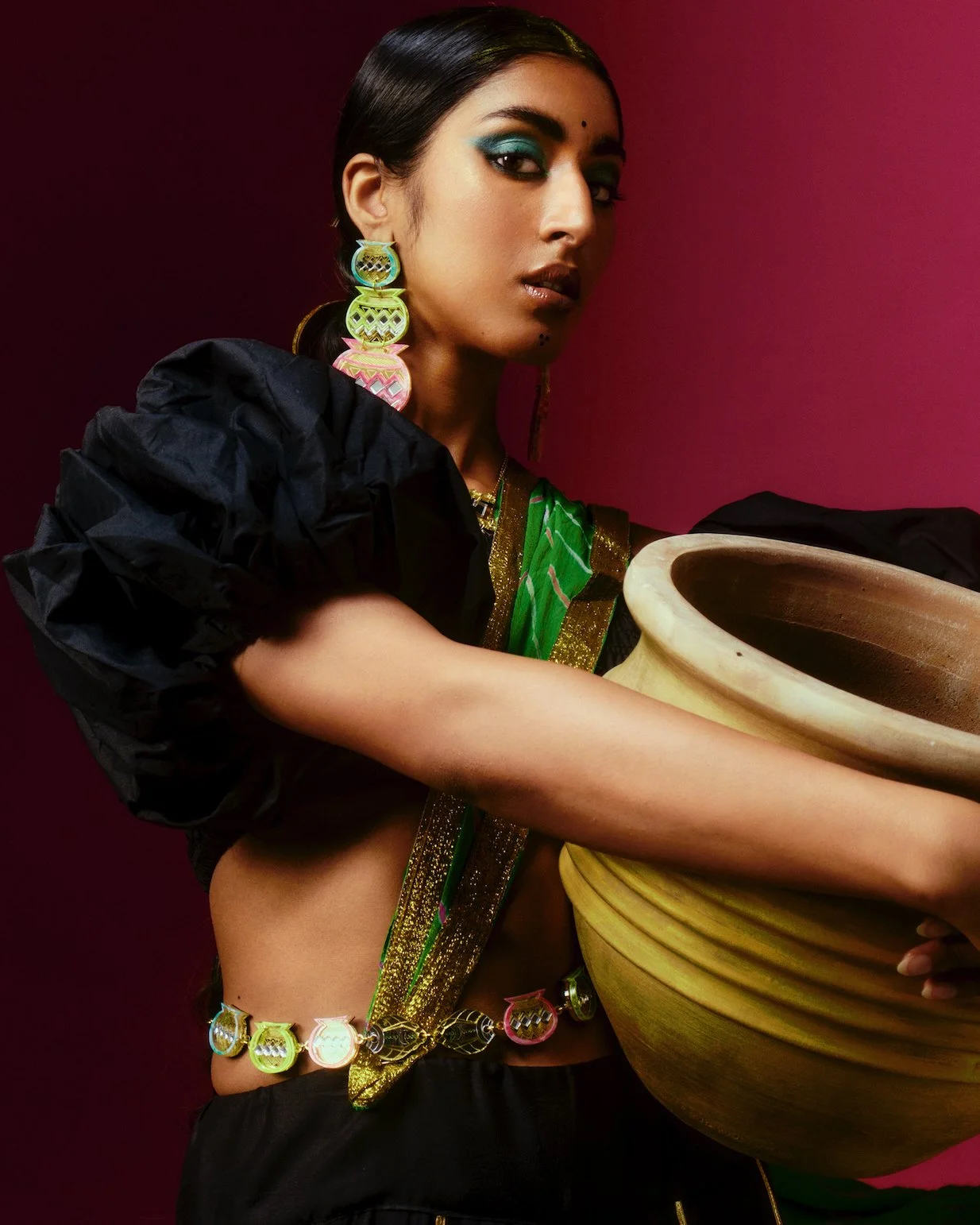 A young woman dressed in puffed-sleeve top in black, matching bottoms, and colorful swathes of textiles and jewelry poses for the camera while holding a ceramic vase and wearing blue eye shadow in front of a bordeaux background.
