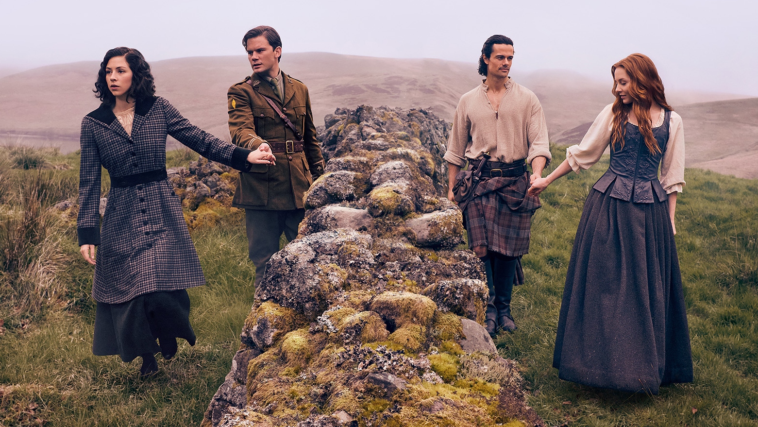 Julia and Henry hold hands on one side of a wall, while Brian and Ellen hold hands on the other side in the Scottish Highlands
