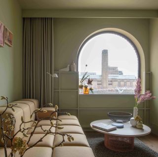 A room in the Locke at Broken Wharf Hotel. Olive green walls with a light brown couch and a round white table take up the entire space. Through a big arched window, we see other buildings.