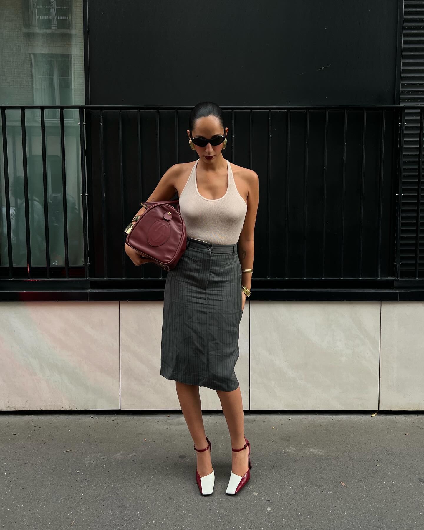 Fashion person Emily Nitta wears a cream halterneck top, a tailored skirt and two tone heel. She is carrying a matching burgundy duffle and oval sunglasses in a recent image from her Instagram.