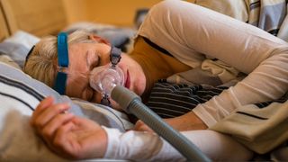 A woman laying on her side in bed wearing a sleep apnea mask and white pyjamas