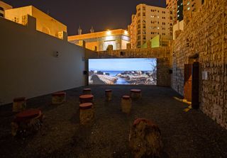 An open walled courtyard styled as an open cinema space, with timber chairs and a big cinematic screen . Photographed at night