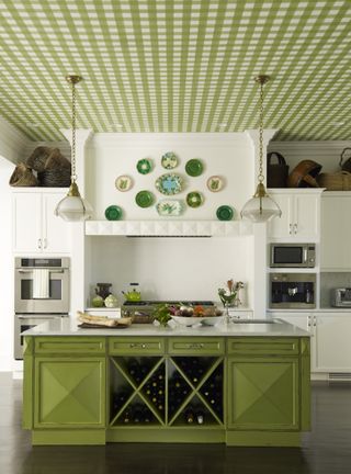 green kitchen with green check ceiling wallpaper, green kitchen island, two vintage pendants, wine storage, hardwood floor, range in fireplace alove, green patterned plates on wall, baskets