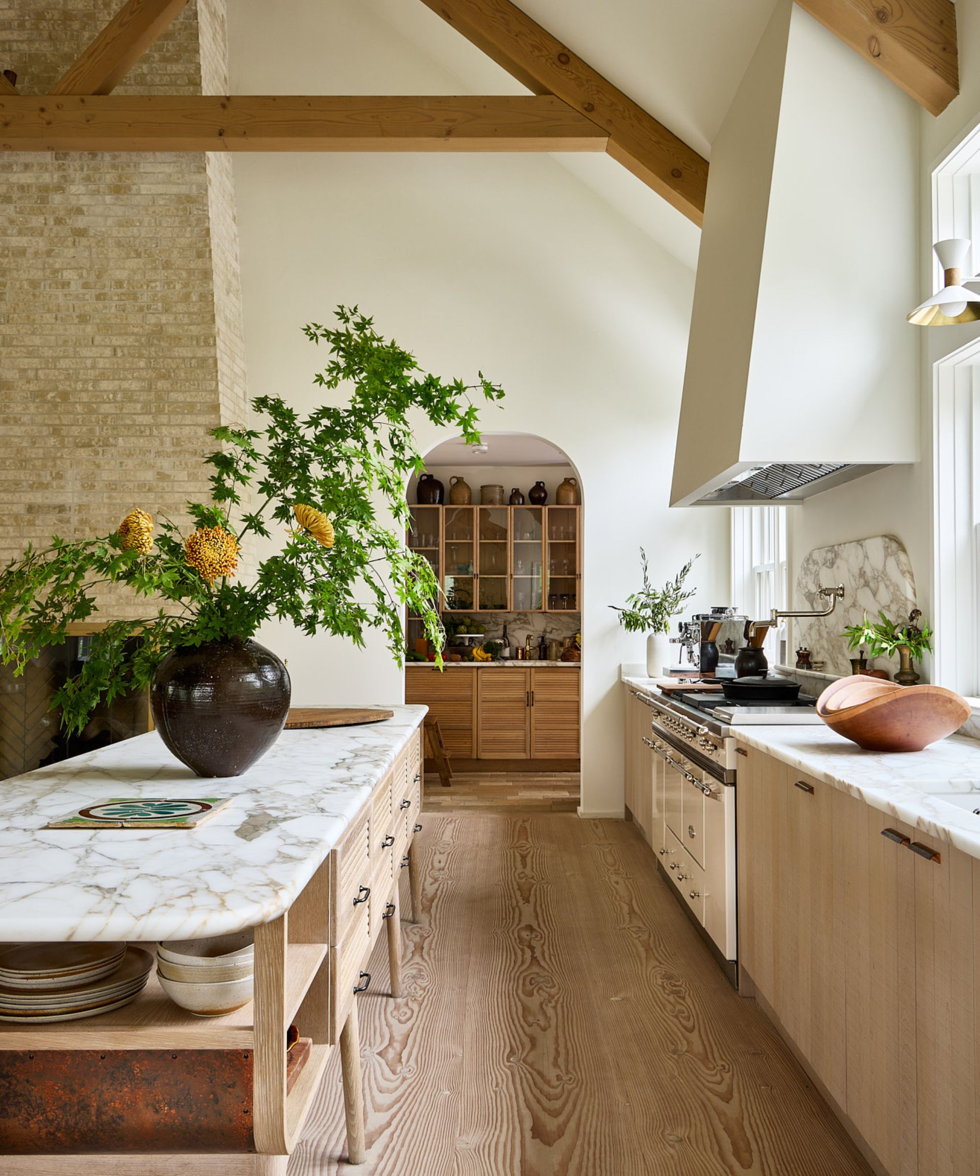 A Coastal Scandinavian kitchen with wooden cabinets, marble countertops, and an arched doorway into the pantry