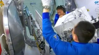 two chinese astronauts in blue flight suits move a white radiation-exposure experiment through the interior of china's tiangong space station
