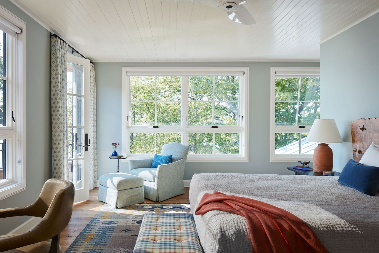 bedroom with pale blue walls double windows all around French doors neutral bed linen organic tree trunk headboard kilim rug Burberry plaid ottoman blue armchair and footstool and white paneled ceiling