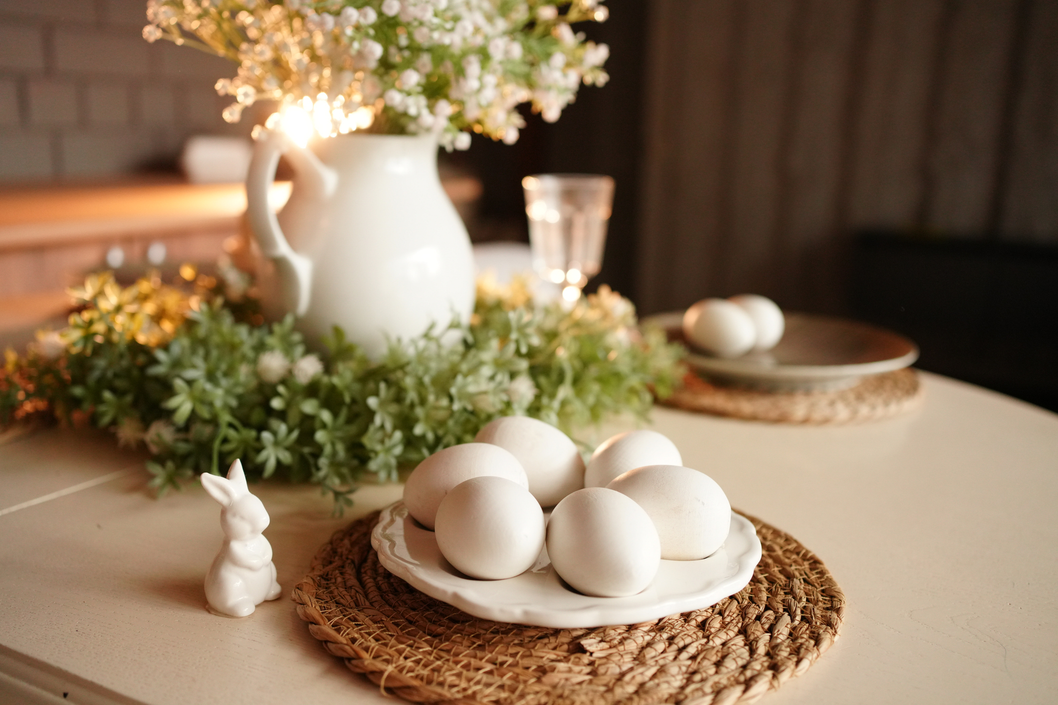 An Easter table spread with white eggs, small, white bunny statues and a white vase with flowers.