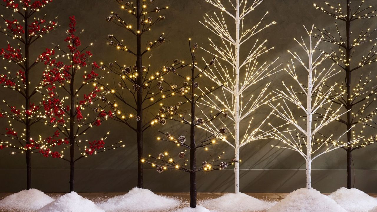 A row of seven artificial Christmas trees that are thin and have branches with lights rather than a full tree. There are little piles of snow beneath the trees