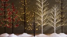 A row of seven artificial Christmas trees that are thin and have branches with lights rather than a full tree. There are little piles of snow beneath the trees