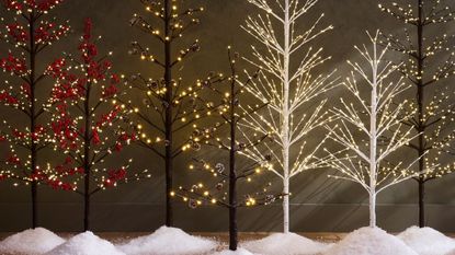 A row of seven artificial Christmas trees that are thin and have branches with lights rather than a full tree. There are little piles of snow beneath the trees