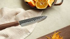 A Japanese knife on a cloth beside a cutting board with orange peel and a bowl of orange slices