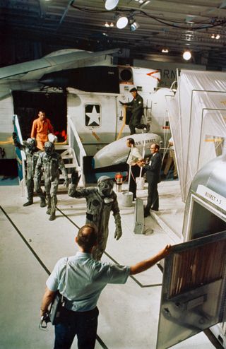 The quarantine procedure began with biological isolation garments, which the Apollo 11 crew donned while leaving the command module and wore until they entered the Mobile Quarantine Facility seen in the right foreground.