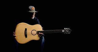 Lloyd Baggs wears a ballcap and spectacles and is pictured in a dark room with the groundbreaking AEG-1 acoustic-electric guitar.