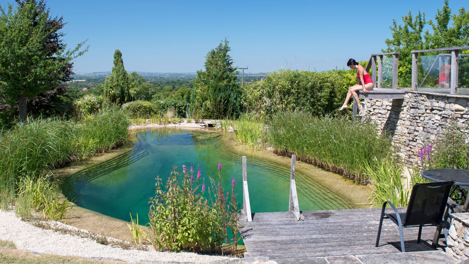 Natural swimming pool surrounded by lush greenery and stone walls, with a woman dipping her toes in from a wooden deck on a sunny summer day.