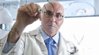 A man in a lab coat and glasses writes equations on a transparent whiteboard