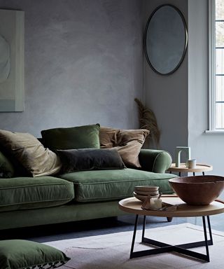 grey living room decorated with a circle mirror with a green sofa and dark pillows and a wooden circled coffee table