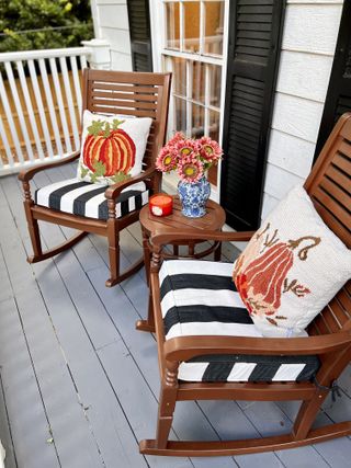 two rocking chairs with fall cushions on front porch, side table with flowers, gray painted wooden boards, black shutters on window
