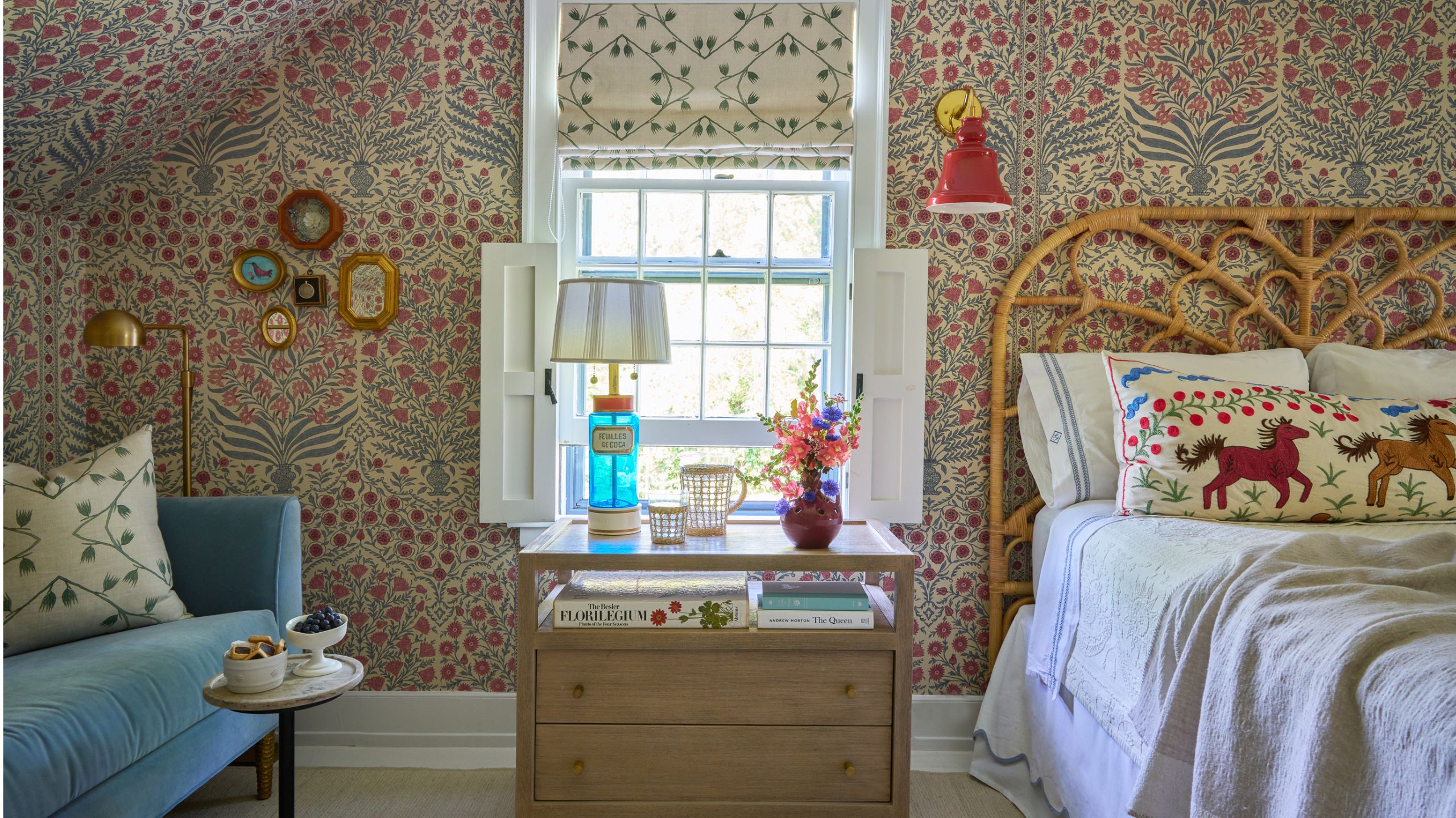 Bedroom with floral wallpaper, a pale blue velvet sofa, a rattan headboard with linen bedding and a folk-style pillow, a nightstand with a vintage lamp and vase of flowers on top, and a window.