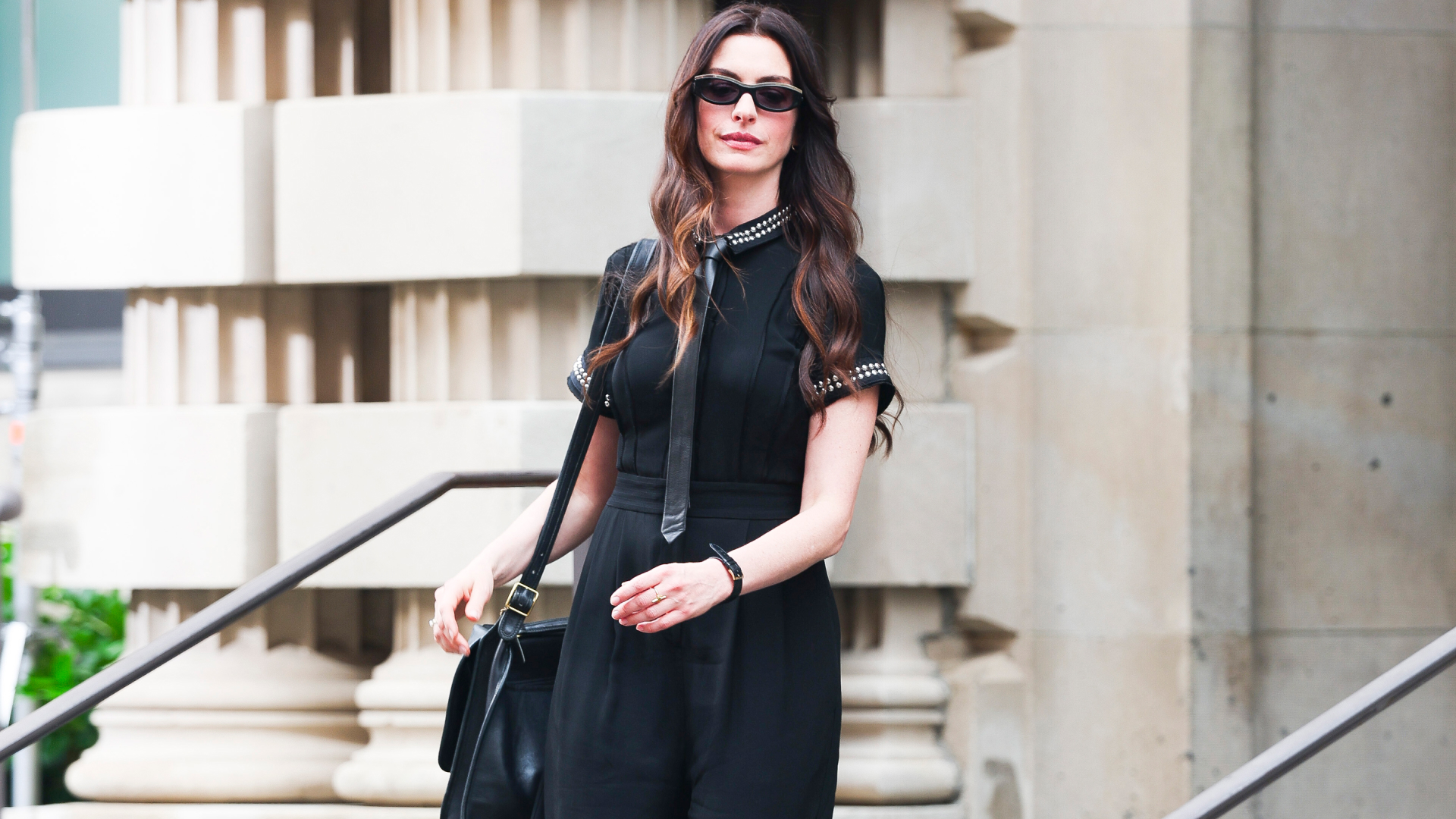 Anne Hathaway wearing a black collared shirt, tie, messenger bag, and sunglasses. She has curls in her brown hair. 
