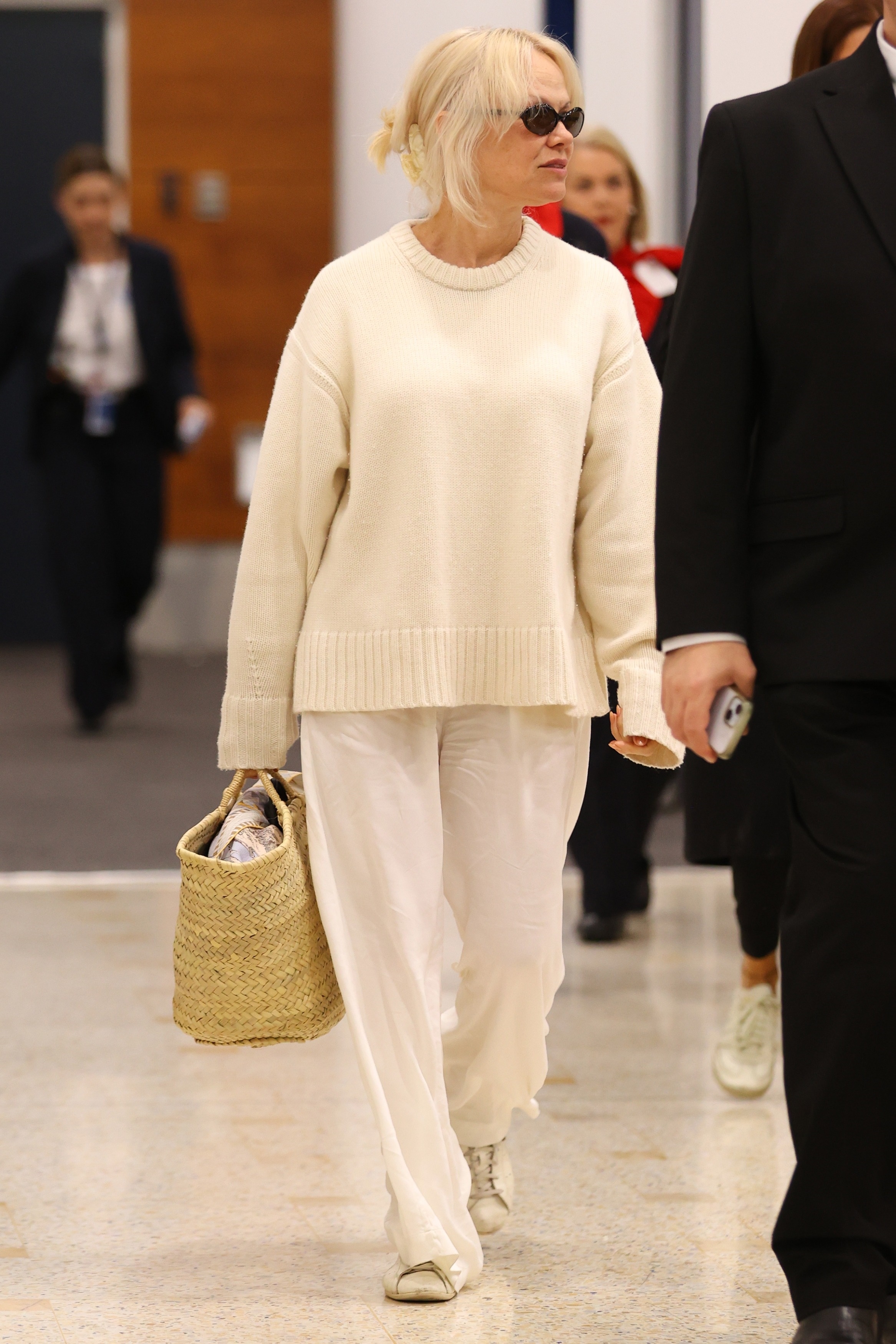 An image of Pamela Anderson wearing a chunky cream knit jumper, linen trousers, white trainers and a basket bag at the airport.